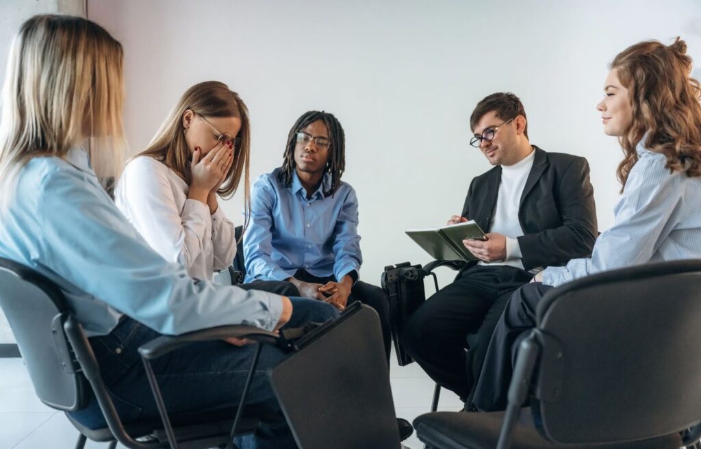 A therapist with a notepad observes as one person cries during a group grief counseling session.