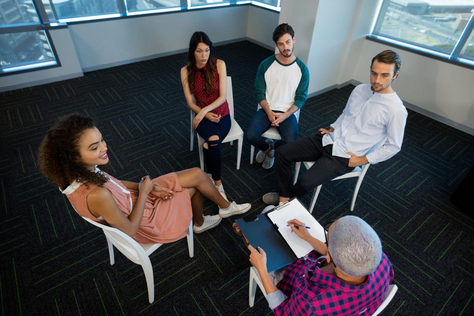 A group of young adults undergo group therapy as their psychologist takes notes.